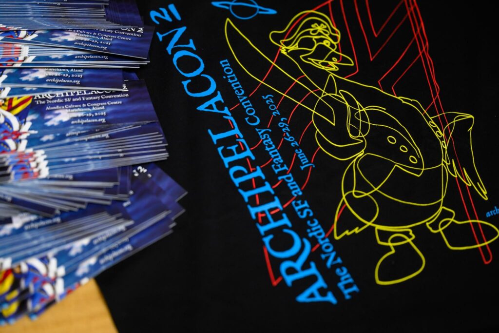 A photo of a black t-shirt with bright blue, yellow, and red text "ARCHIPELACON 2" on it. Underneath the text is a smaller text, "The Nordic SF and Fantasy Convention, June 26–29, 2025". The text is set over an image of a cartoon pirate swan, designed in yellow lines. In the foreground of the image is a stack of bookmarks for the event.