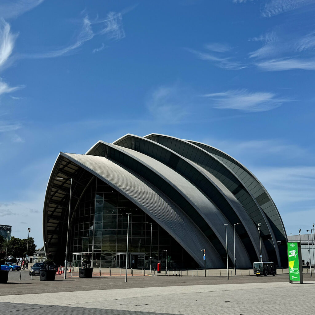 The “Armadillo”, a modern, multi-arched, silver building with a glass front, against a clear blue sky.